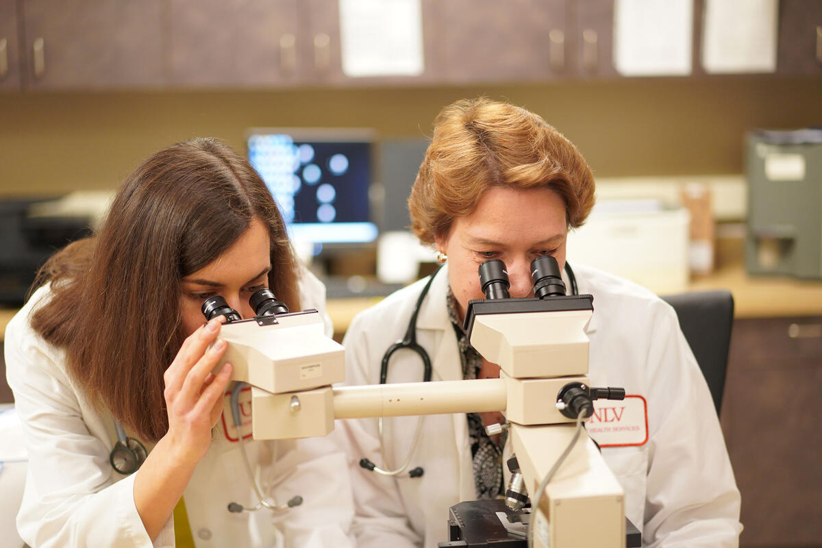 Two women in lab coats looking through their own microscope