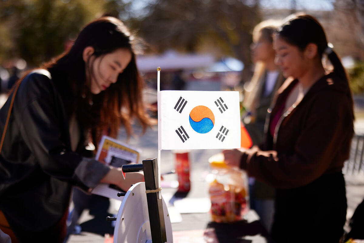 Two people at a table with a South Korean flag displayed prominently in the foreground. One person is out of focus, interacting with a table, while another person is seen in the background.