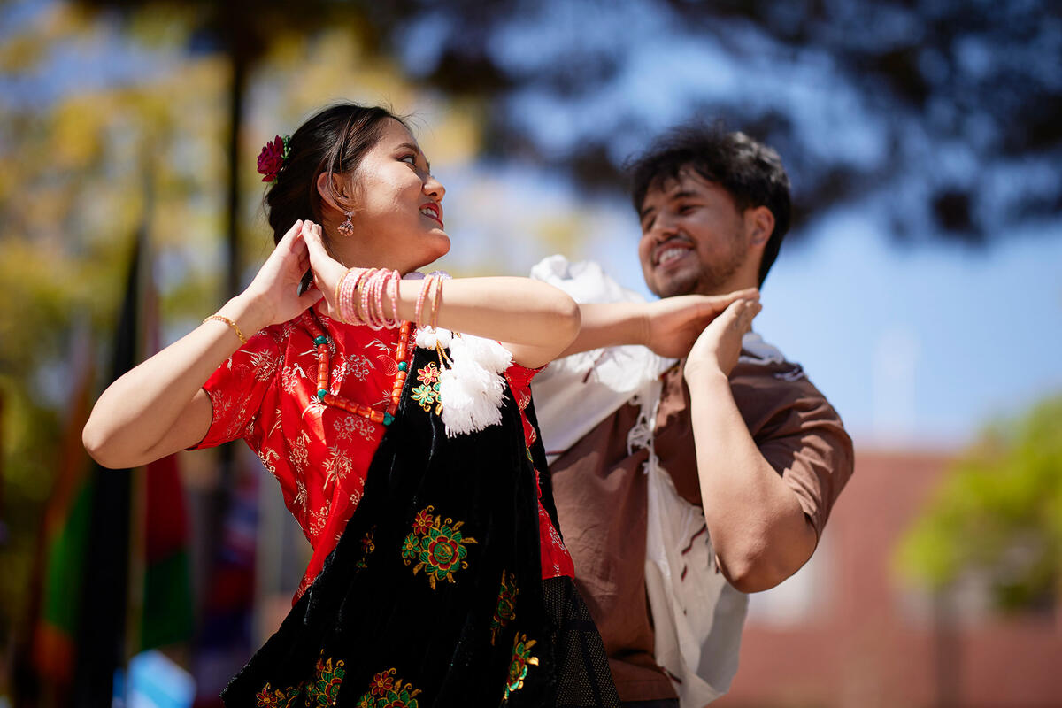 Two individuals share a joyful moment while dancing together outdoors, with the woman dressed in a floral dress, and the man in a brown shirt. Colorful flags can be seen behind them.