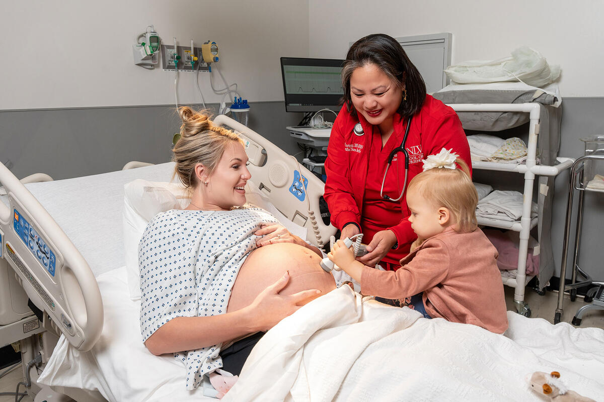 A pregnant woman lies in a hospital bed smiling while a young girl holds a fetal monitor to the woman’s belly with the help of a nurse in a red UNLV School of Nursing uniform