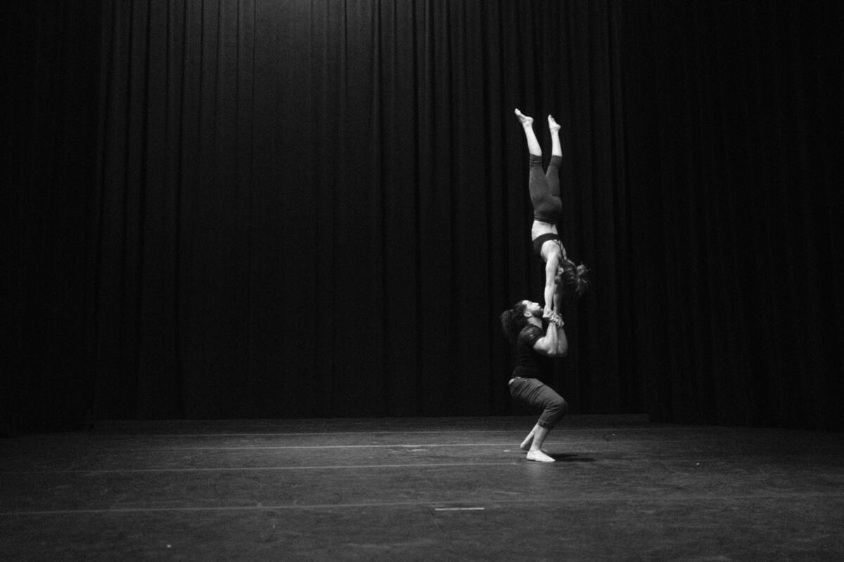 male acrobat lifts female acrobat into a handstand on dark stage