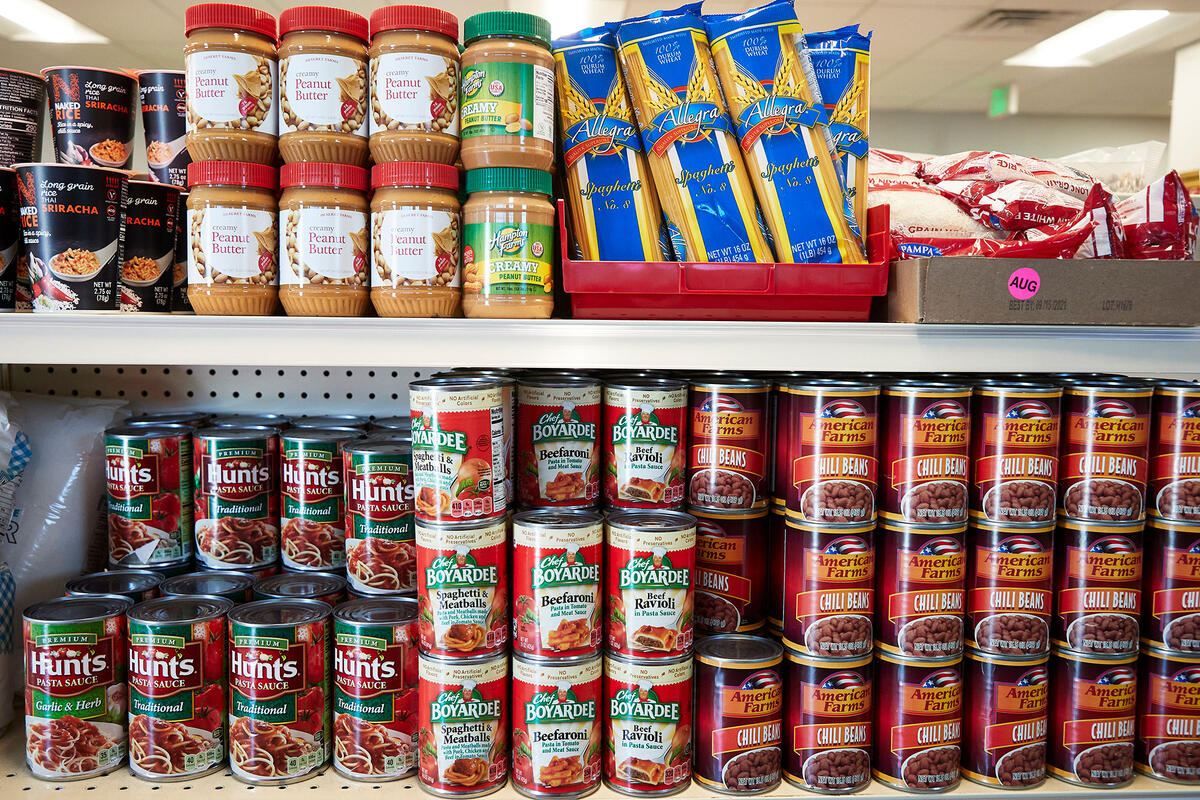 A shelf in a food pantry with various items including cans of pasta sauce, chili beans, Chef Boyardee meals, jars of peanut butter, and packs of spaghetti noodles.