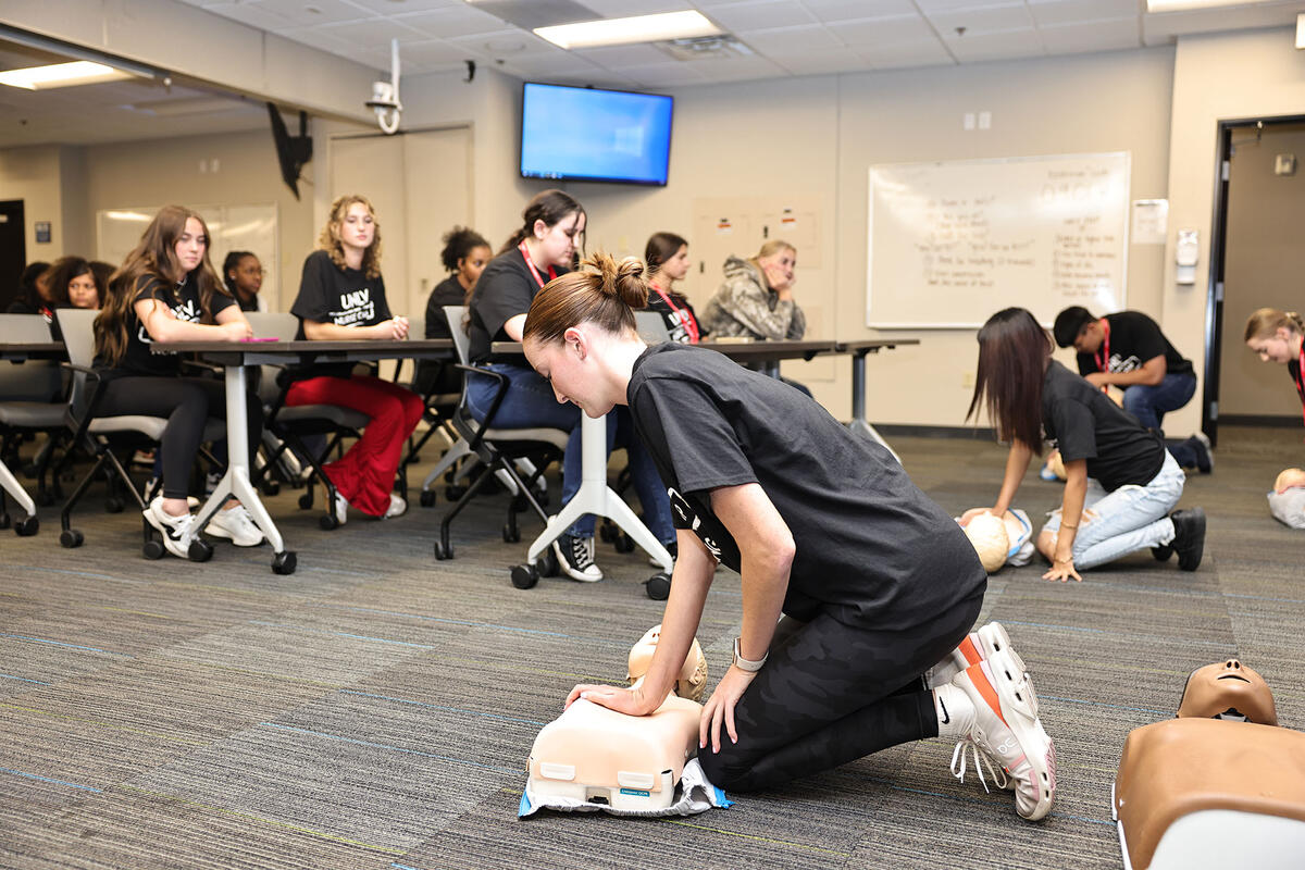 Participants in black "UNLV Nurse Camp" shirts practice CPR techniques on training manikins in a classroom setting, while others observe from desks in the background.