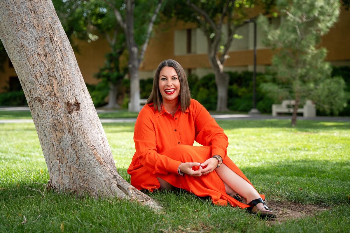 White woman with shoulder length brown hair wearing a bright orange dress sits on grass under a tree on UNLV's campus