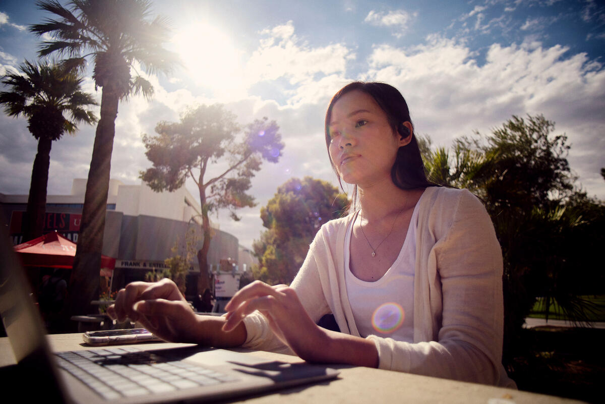 A student studies at a laptop in an outdoor seating area in front of palm trees as sun shines through the clouds