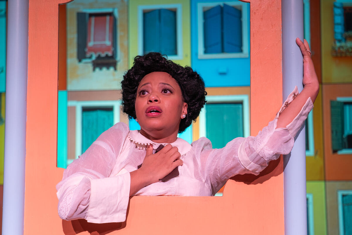 A woman looking out to the audience in a colorful setting. Her left hand is held out to the side as she grasps an object close to her chest with her right hand