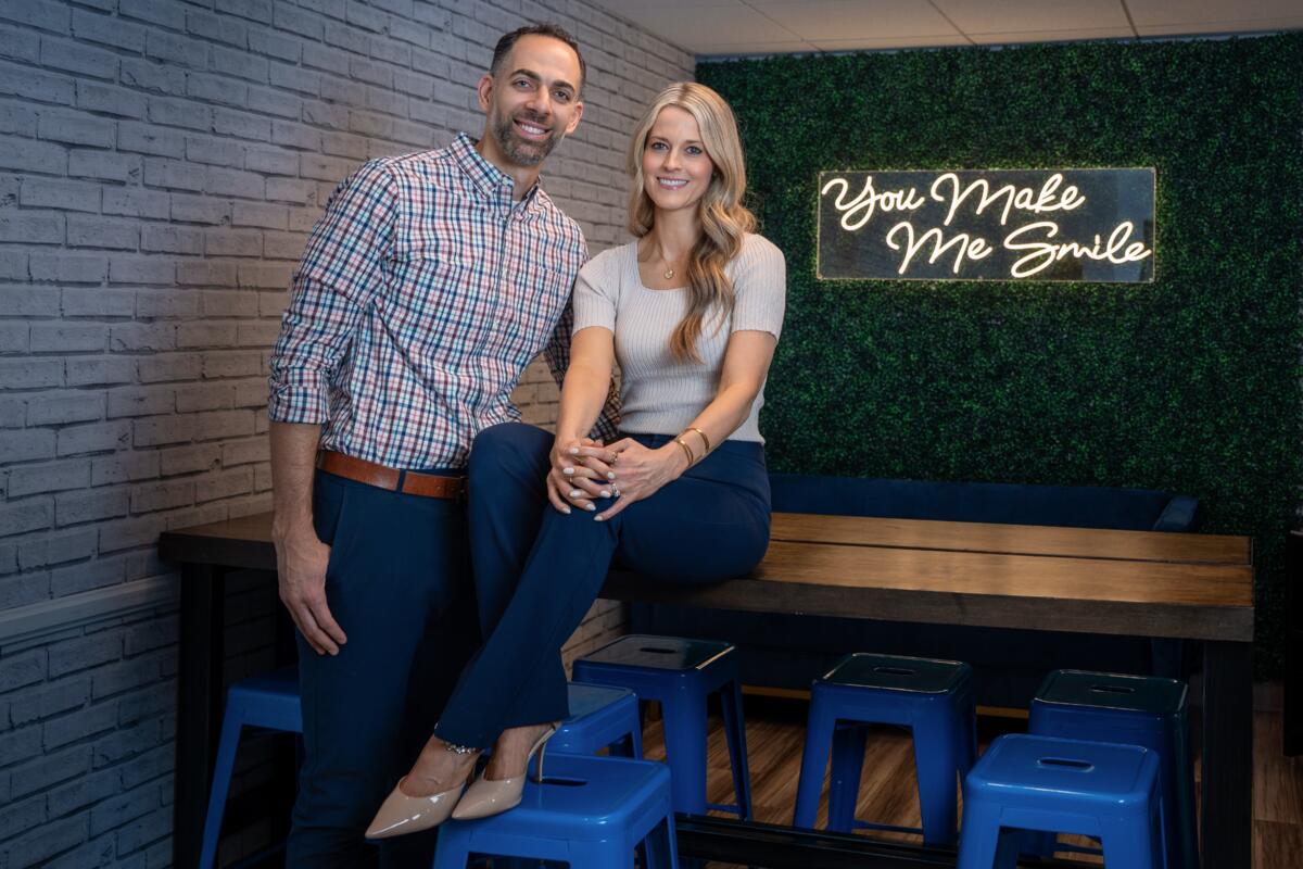 A white woman with long blonde hair and wearing a cream-colored top, blue pants and beige high-heel shoes sits on a dark wood table with her hands clasped around her knee as a man with thinning dark hair, plaid shirt, brown belt and dark pants leans in from her right. Both are in front of a lighted sign with the words You Make Me Smile in cursive script