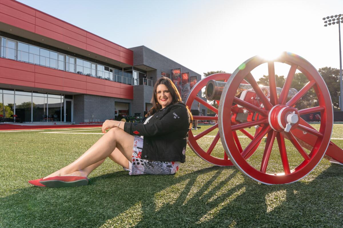 White woman with long brown hair wearing a dark jacket with Class of 2008 emblem and white, black, and red shorts and red shoes poses while sitting on grass with her back against the wheel of a red cannon outside the UNLV football complex