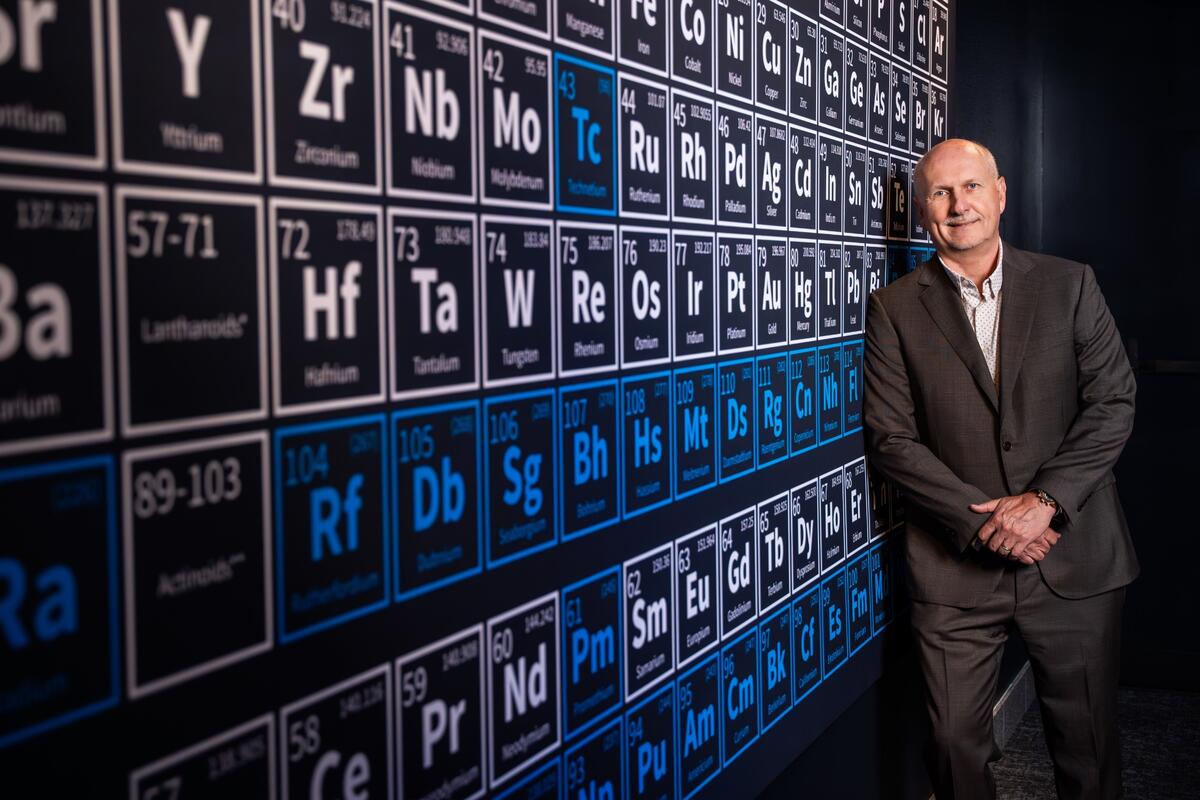 Bald white man in a brown suit standing next to an oversize periodic elements chart