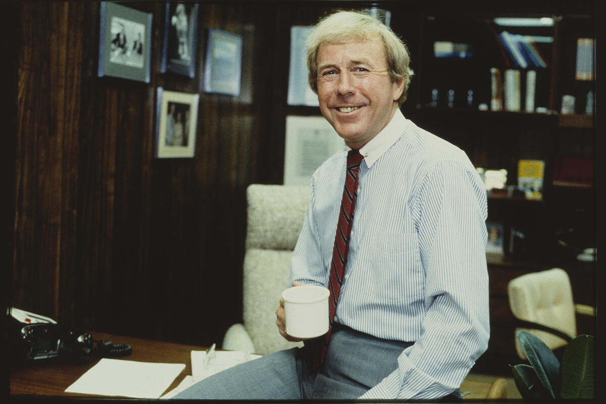 archive photo of man in tie holding coffee cup and leaning on office desk