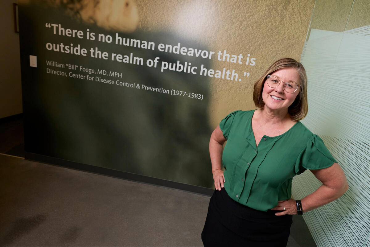 A portrait of Kelly Webber. Behind her, a quote on the wall reads, &quot;There is no human endeavor that is outside the realm of public health.&quot;