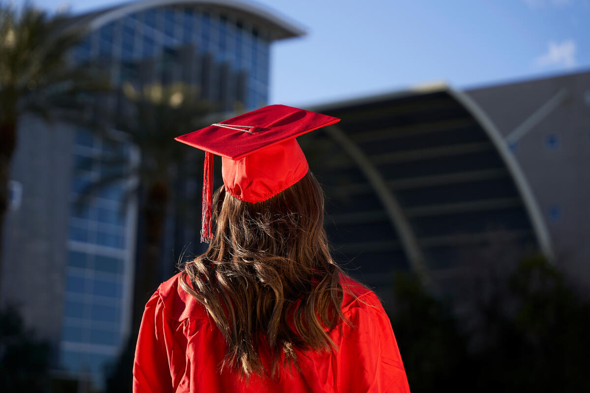a female student with dark hair and wearing a red cap and gown is shown from the rear, facing Lied Library