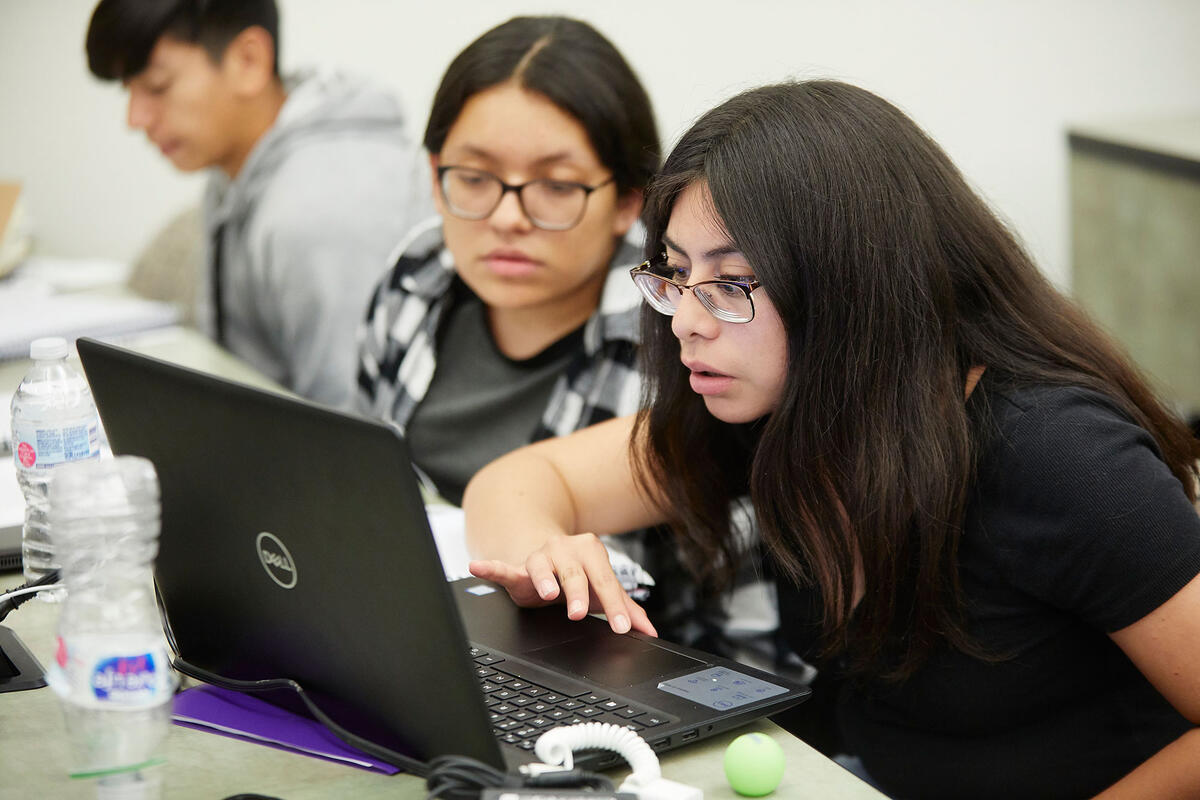 Two students sit closely together at a desk, focused on a laptop screen.