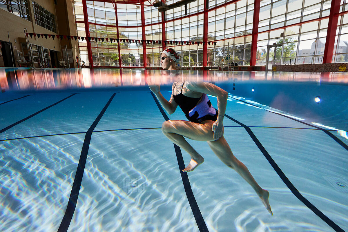 Underwater view of an adult swimmer wearing a flotation belt and swim cap, running along the lane lines in an indoor pool.