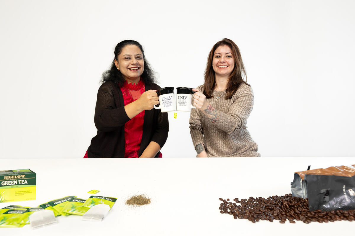 Two women raise two mugs in a toast pose. Green tea bags are arranged on one side of the table, and coffee beans on the other side.