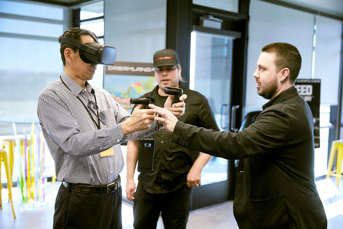 A man wearing a VR headset and holding controllers while two other men observe
