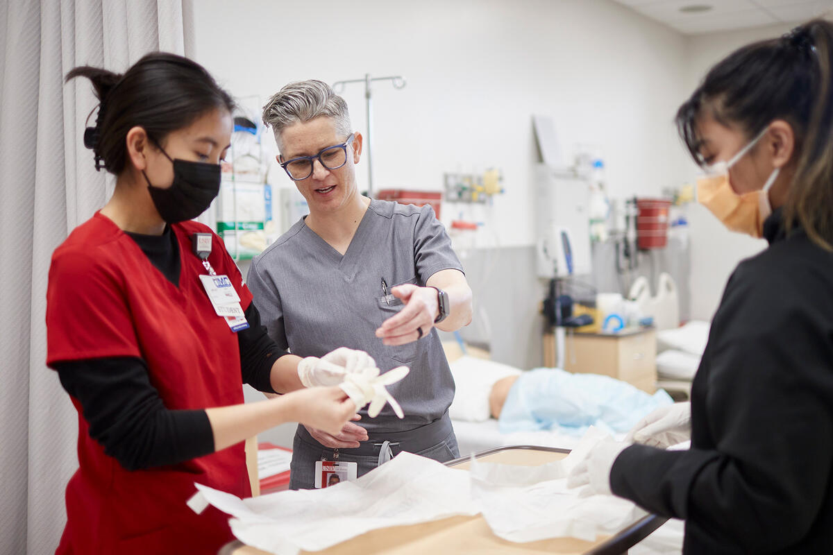 An instructor in gray scrubs demonstrates a clinical procedure to two masked students at a bedside.