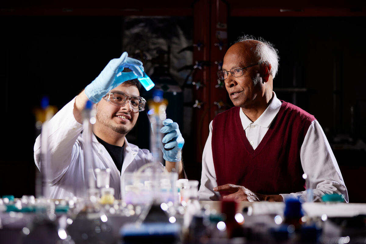 A student wearing safety goggles and gloves holding a glowing blue liquid sample while a researcher observes.