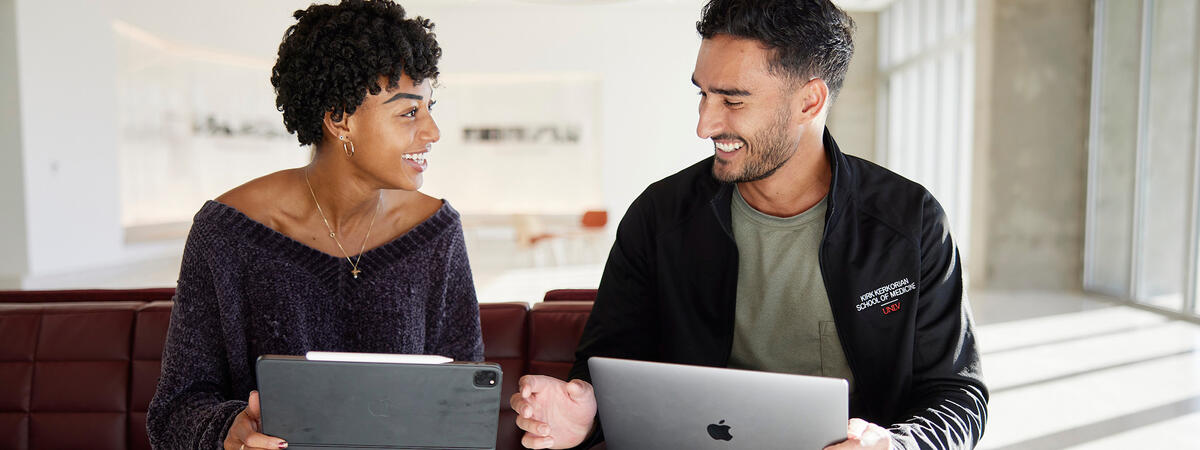 Two people sitting on a red bench, smiling at each other while holding a tablet and a laptop.