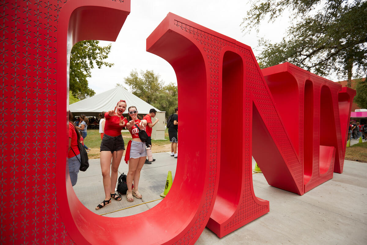 Two female students in shorts and red tops make the LV sign with their hands while standing behind a red "U" at the on campus UNLV sculpture