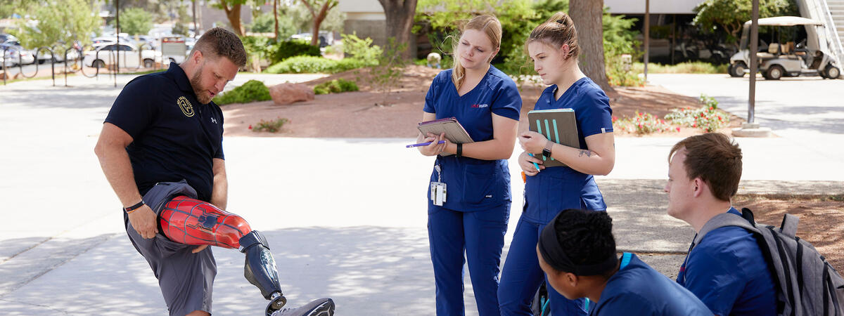 A man showcases a lower-limb prosthetic while standing outdoors as a small group of nursing students in blue scrubs observe and take notes.