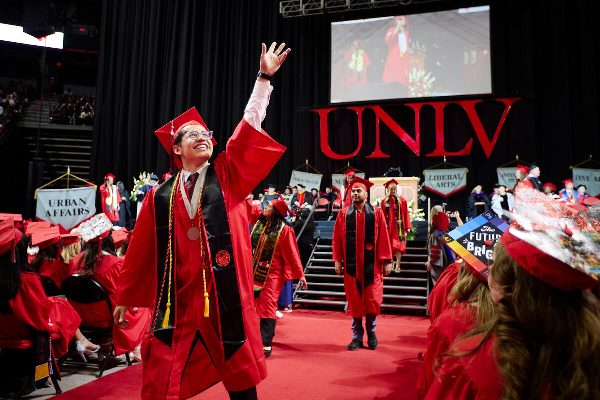 Graduate in red cap and gown waves while walking across the stage.