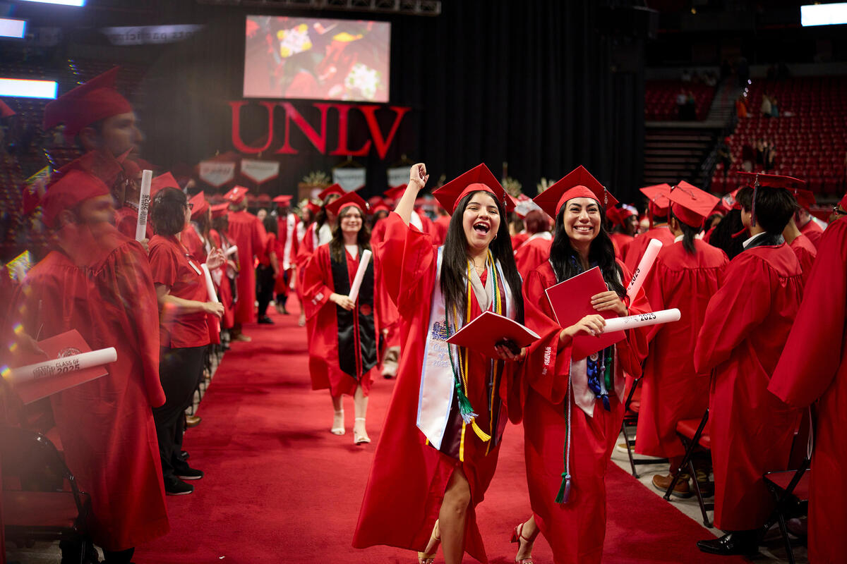 Two graduates in red caps and gowns walk down an aisle while holding a diploma in their hands. 