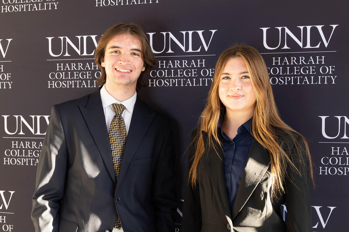 Two people in business attire standing in front of a UNLV Harrah College of Hospitality backdrop.
