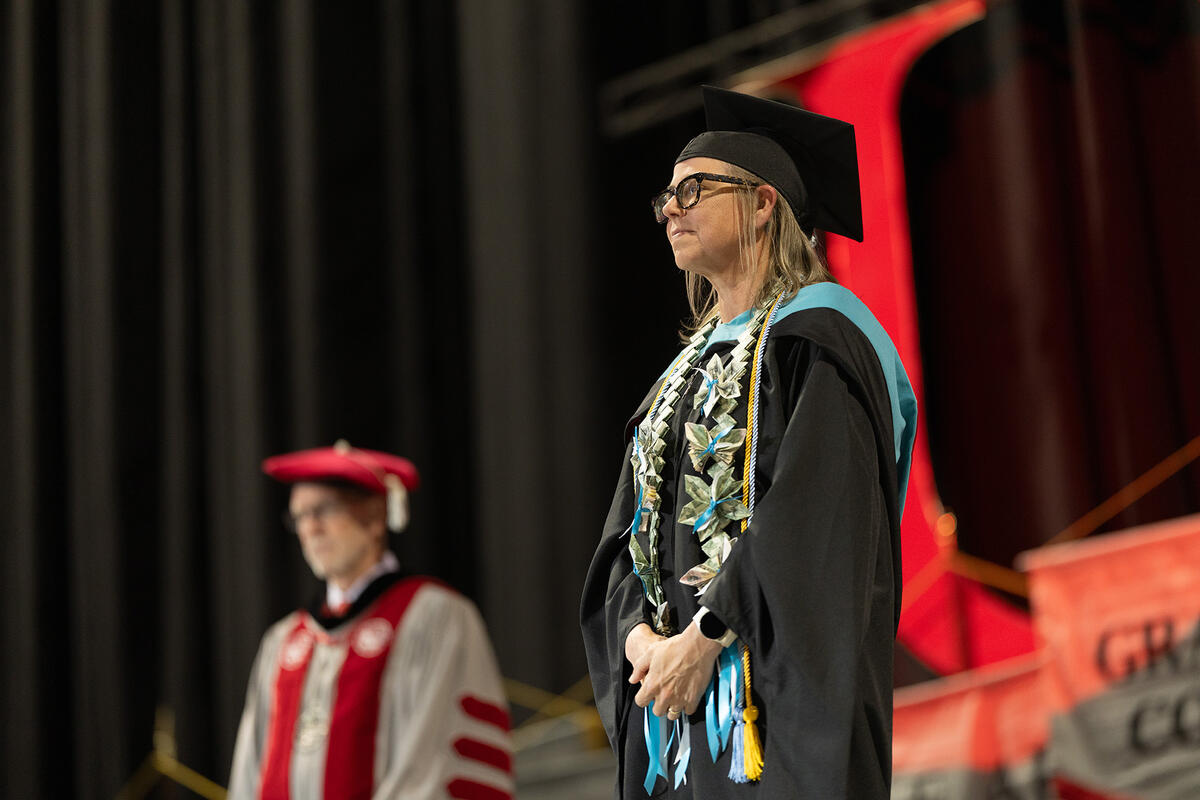 A graduate wearing a cap, gown, and honor cords stands on stage