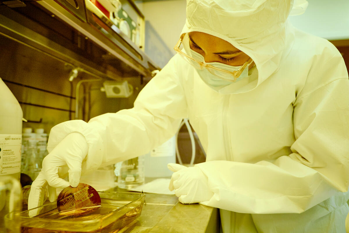 A person in a disposable white hazmat suit pinching a maroon flat circular chip into a liquid