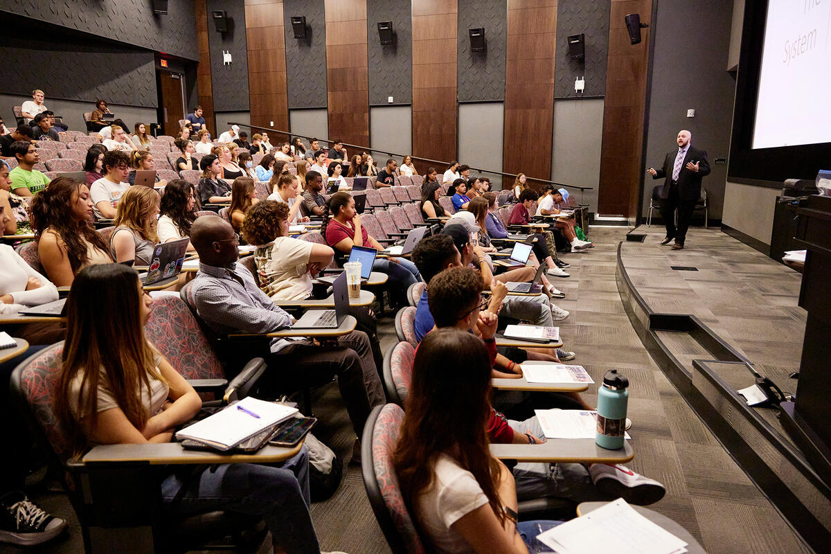 Professor presenting to a lecture hall filled with students seated with laptops and notebooks.
