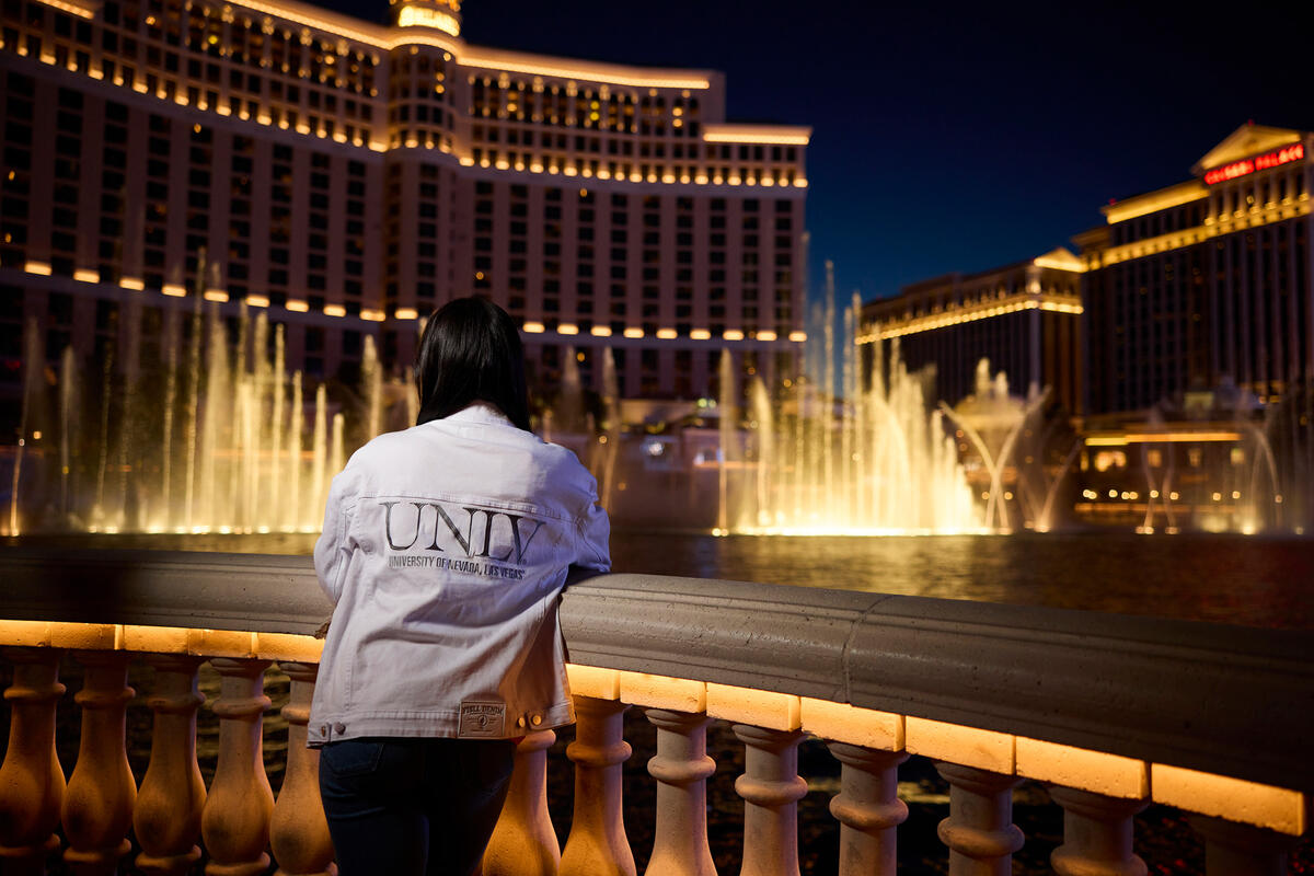 A woman wearing a white demin jacket with "UNLV" on the back standing in front of the Fountains of Bellagio