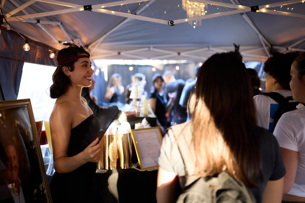 A person in vintage-inspired attire smiles while speaking with attendees at an event booth