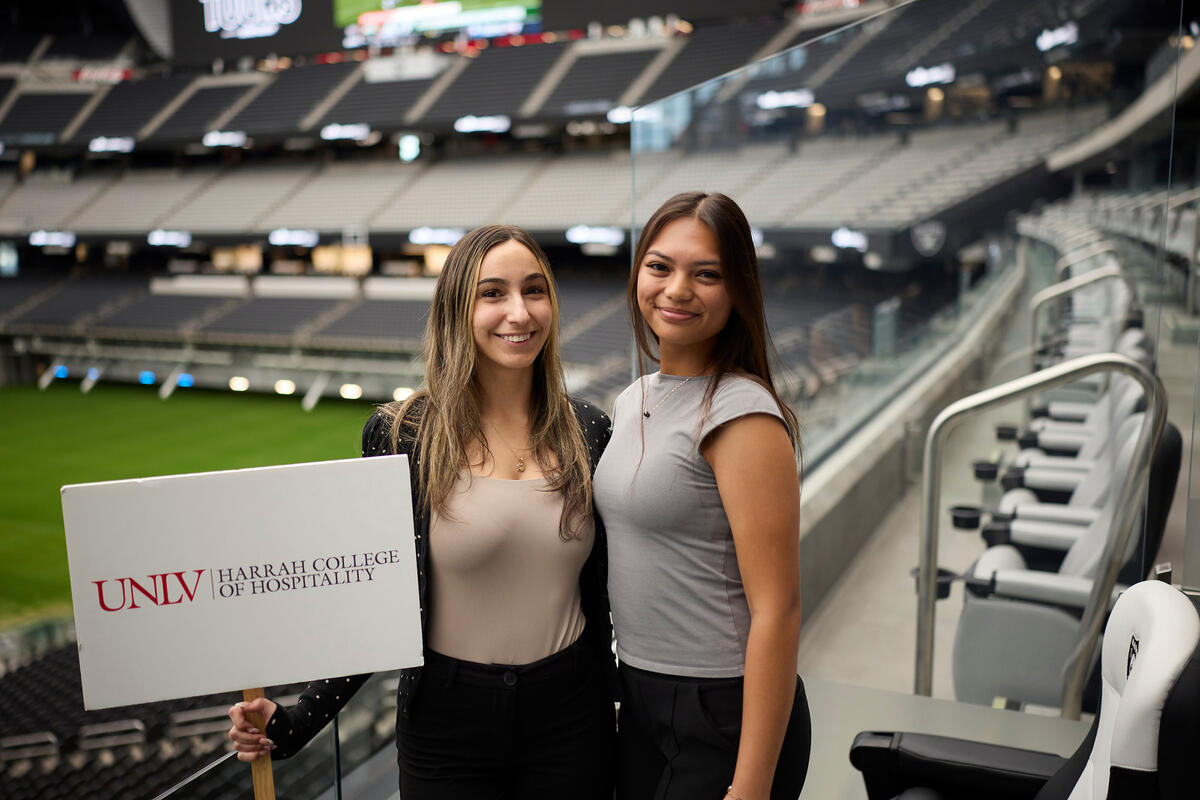 Two students smiles while standing inside of Allegiant stadium, one holding a sign reading "UNLV Harrah College of Hospitality"