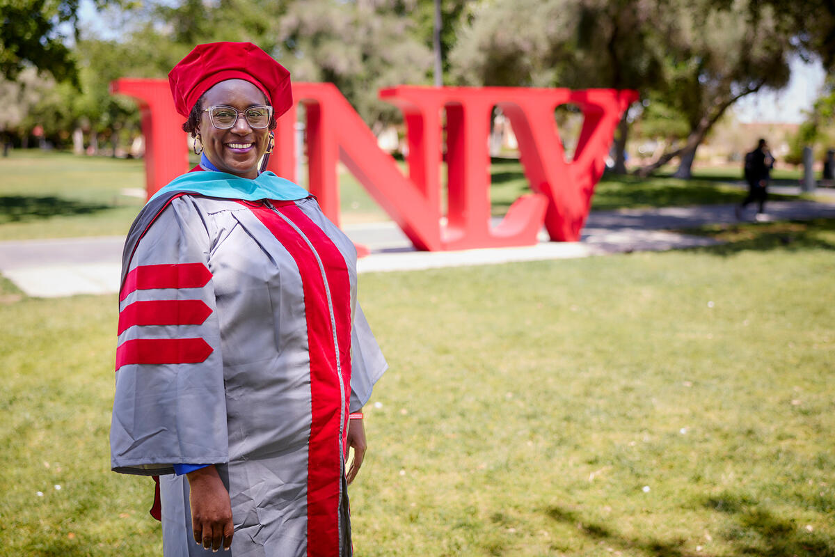 A graduate standing on campus lawn in front of large red U-N-L-V letters
