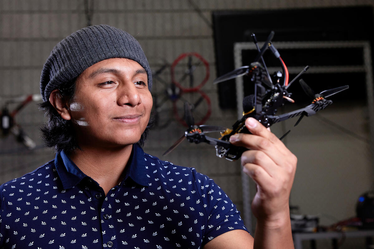 A man smiling in a navy blue polo with white leaves and a gray beanie holding a drone in his left hand