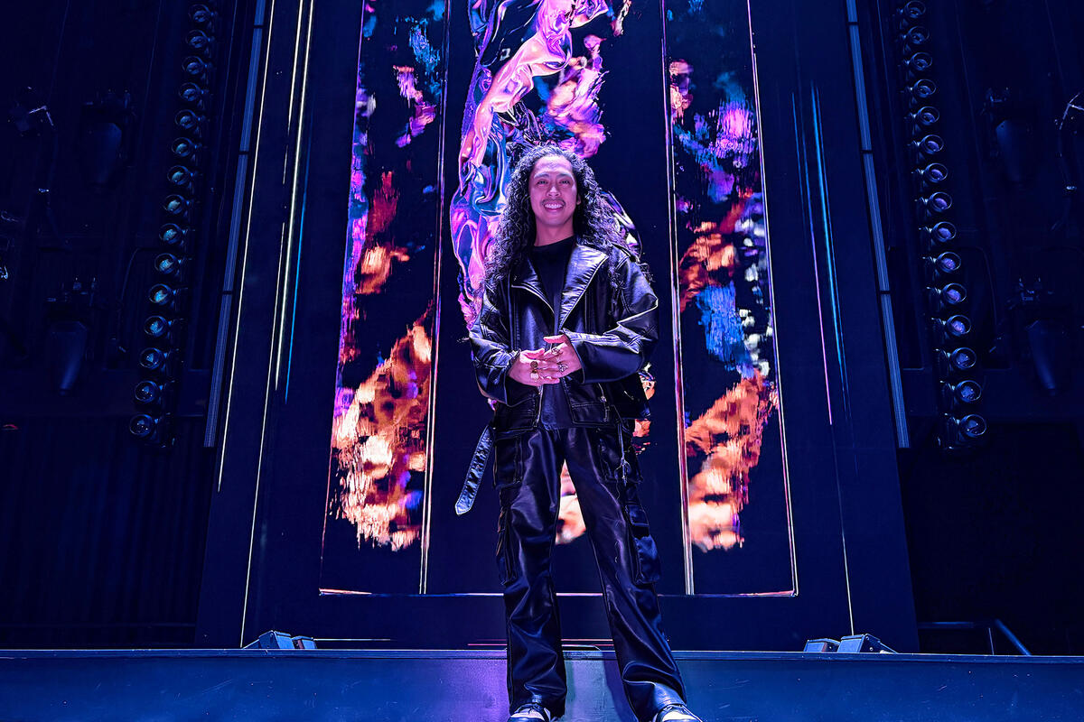 A person wearing a black leather jacket stands on a stage in front of large, colorful digital display panels