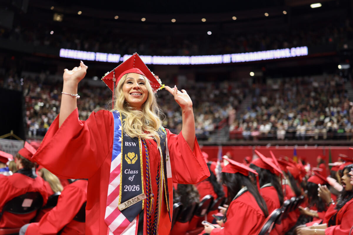 A student in a cap and gown with both hands raised as she waves towards the crowd