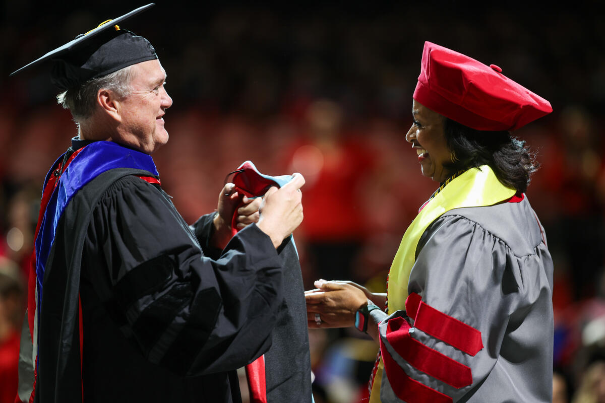 1 student in a black cap and gown holding out a stole to a student in a red and gray doctoral cap and gown