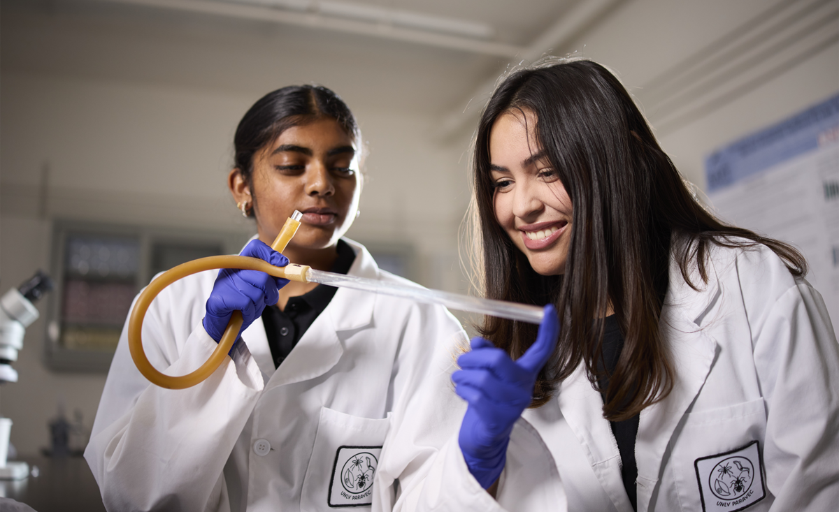 2 women in white coats and dark blue gloves looking at a tube