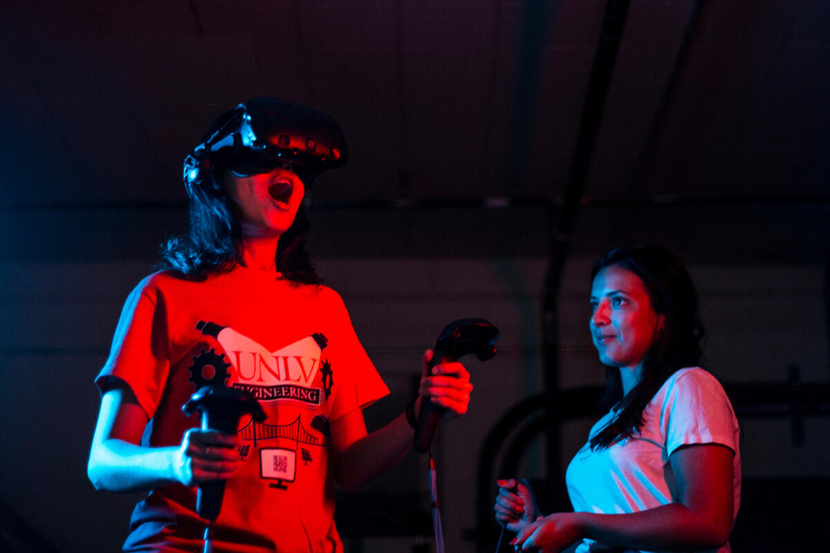 A student in a dark room with a VR headset and a woman standing behind her