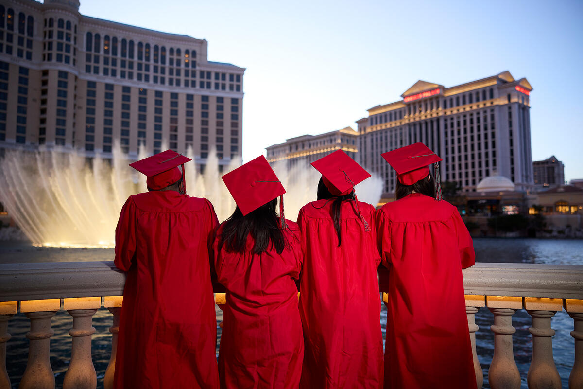 Four graduates in red caps and gowns stand with their backs, overlooking the Bellagio fountain show.