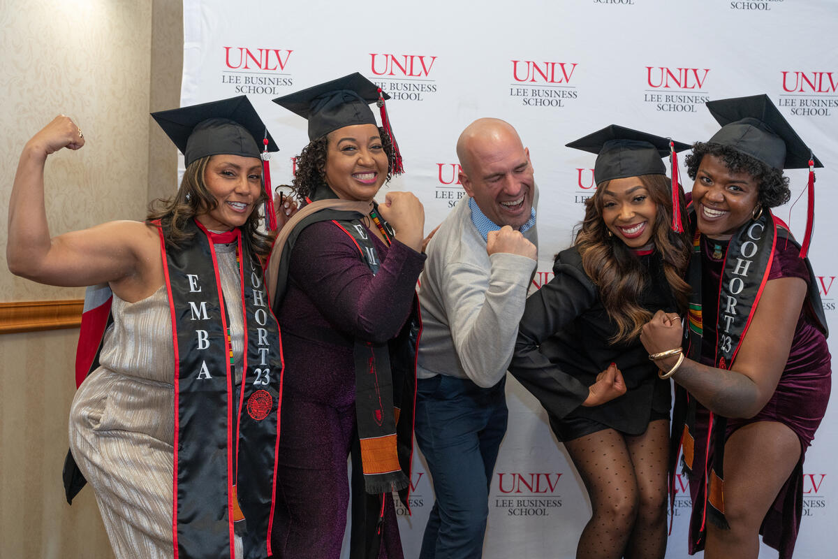 4 graduates and a staff member posing in front of a backdrop