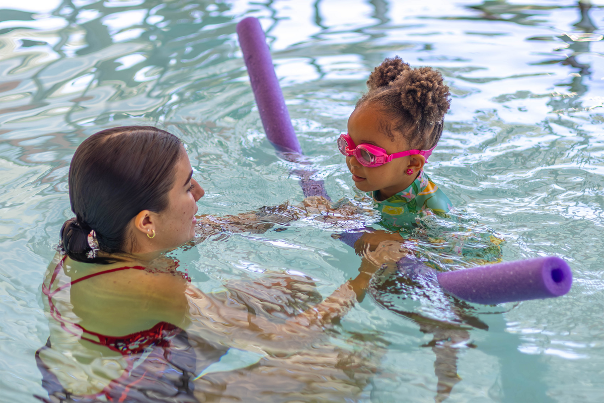 A student lifeguard helping a child with a purple pool noodle swim across the pool