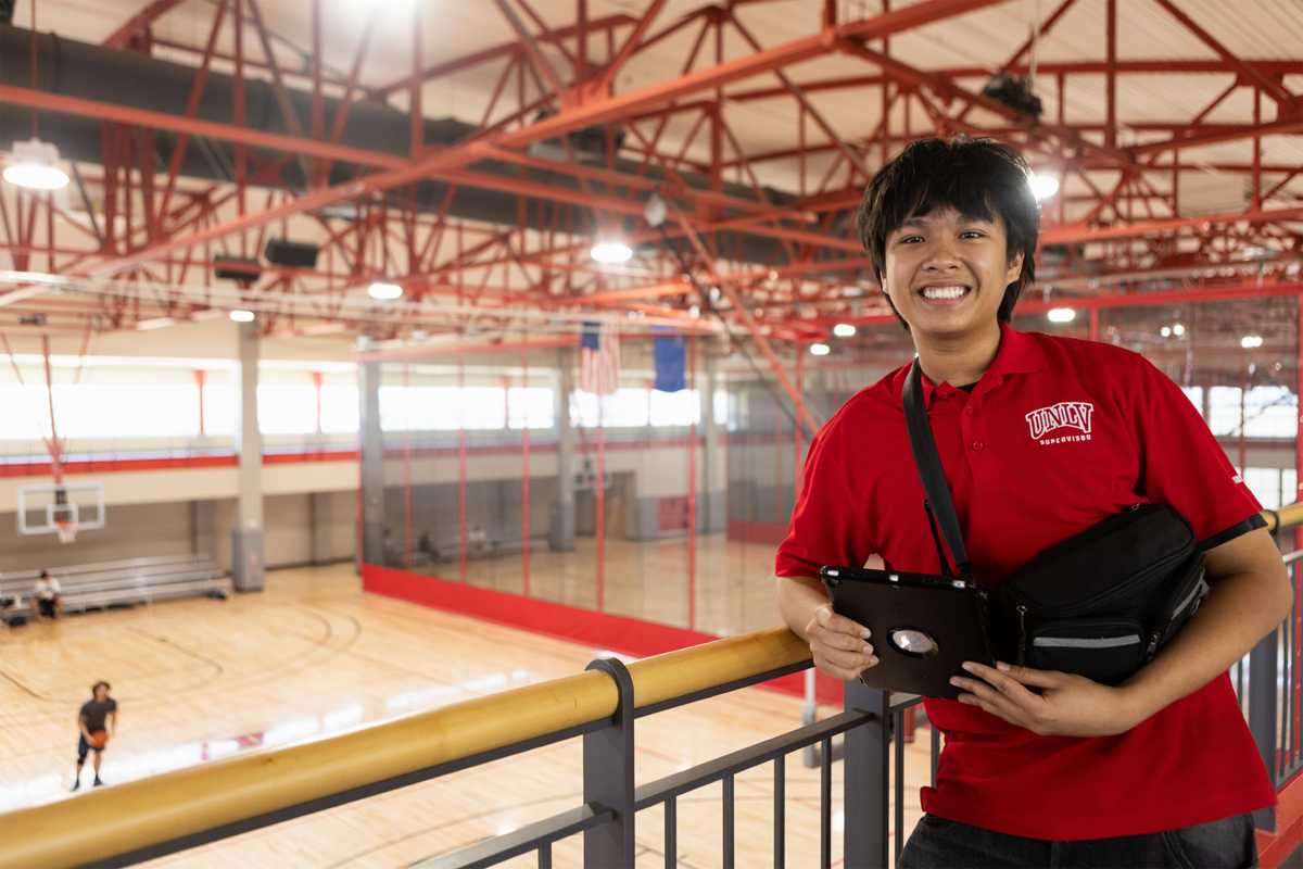 A student worker leaning against a gray railing over the U-N-L-V Student Recreation and Wellness Center's Gym