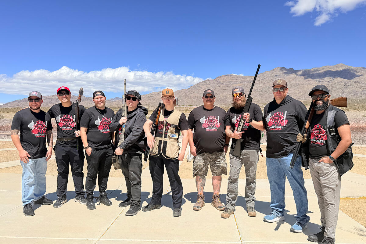 Nine people wearing UNLV Corporate Challenge t-shirts pose with shotguns at an outdoor shooting range