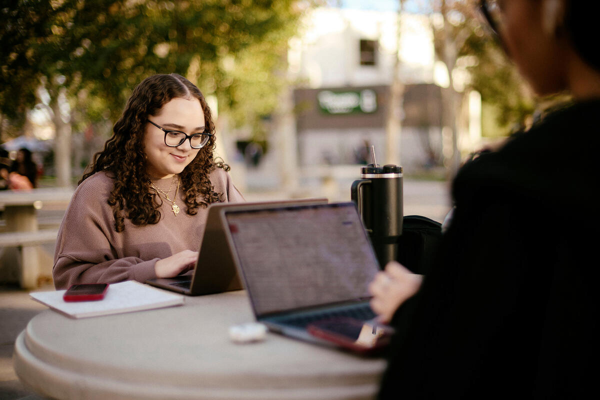 A student wearing glasses and another student in the foreground works on a laptop outside at a table.