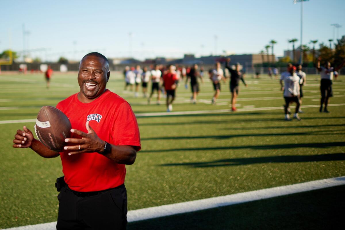 Black man wearing black pants and a red T-shirt with a UNLV logo smiles wide while flipping a football to himself while in the background blurred out players jog on a football field on a sunny day