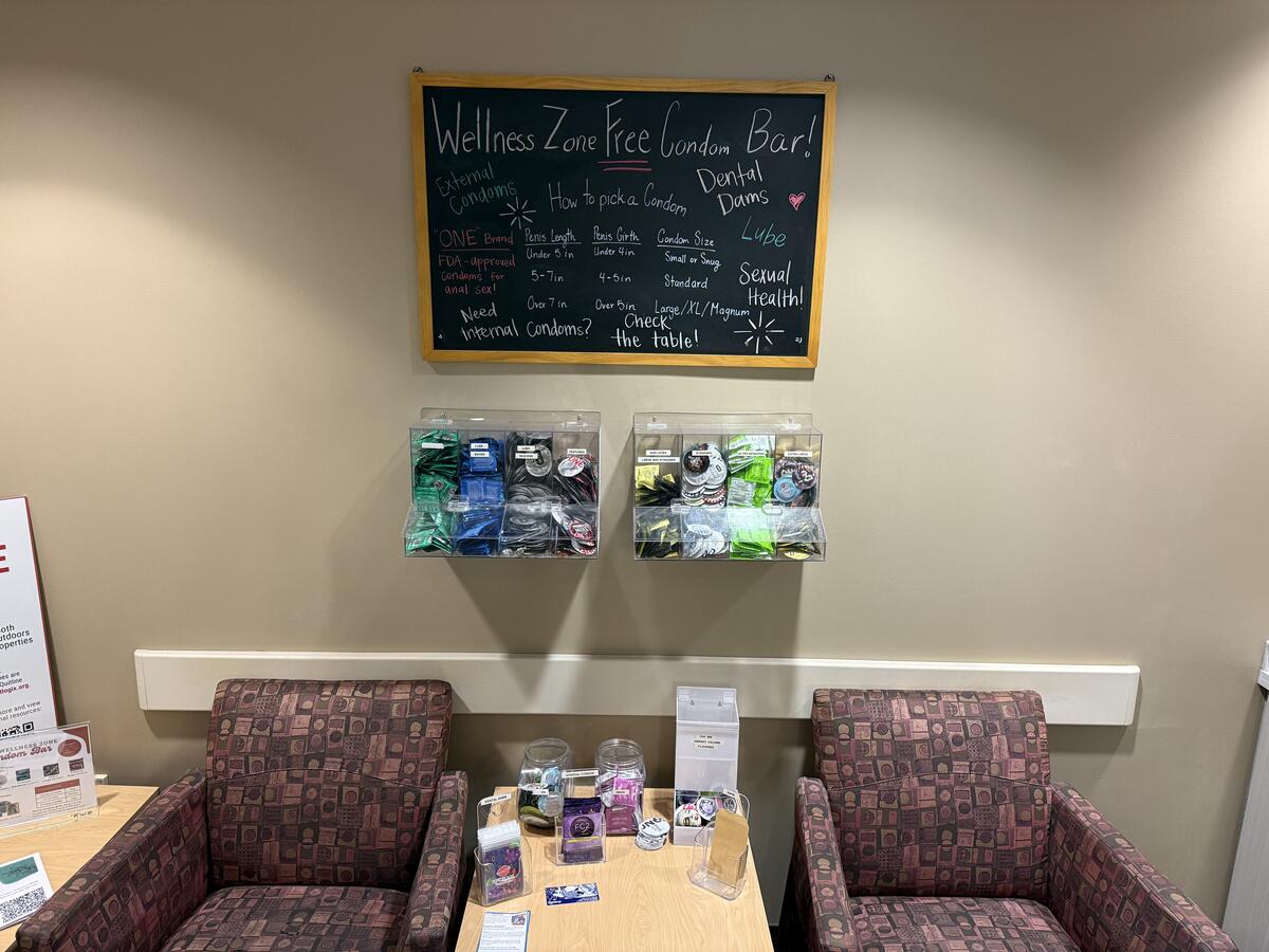 The wall of a lobby with 2 chairs and a chalkboard that says "Wellness Zone Free Condom Bar" in the background