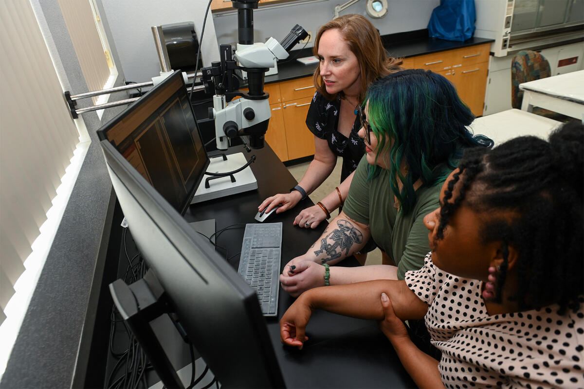 A professor seated by a computer with two students observing